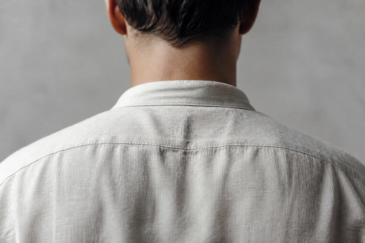 Nuque et épaules d&rsquo;un adulte vus de dos en chemise de lin claire, lumière naturelle latérale, cadrage serré